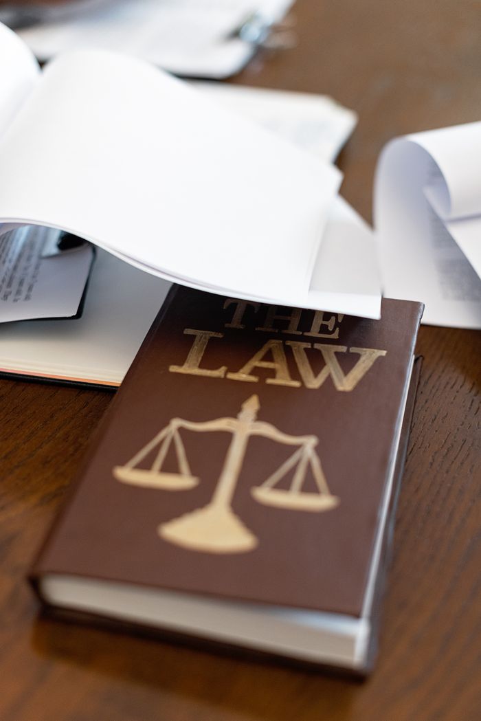 services-img-01 Close-up of a law book with scales icon on a wooden desk covered in papers.